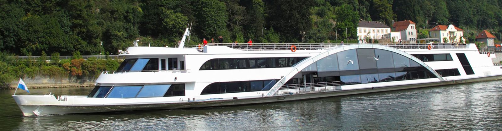 Modern river cruise ship with a glass-roofed lounge sailing past a wooded bank