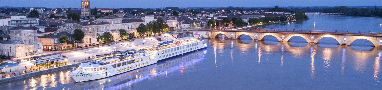 River cruise ship lit up at dusk beside an old stone bridge